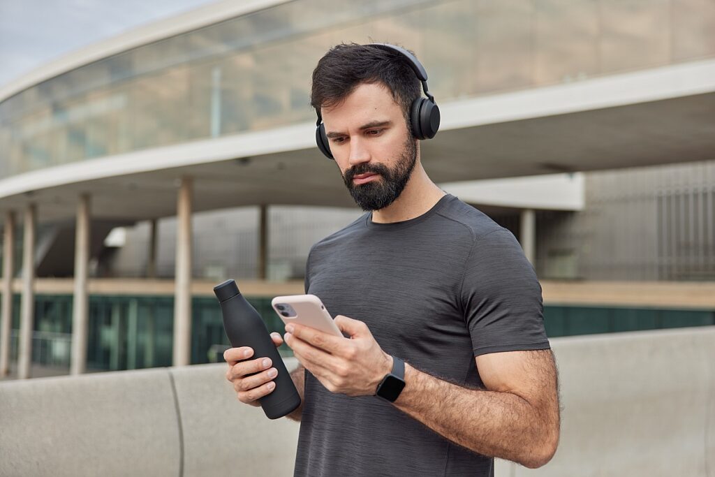 muscular athletic man looks smartphone screen downloads songs playlist training holds bottle with fresh water wears casual t shirt has regular workout poses outdoors healthy lifestyle