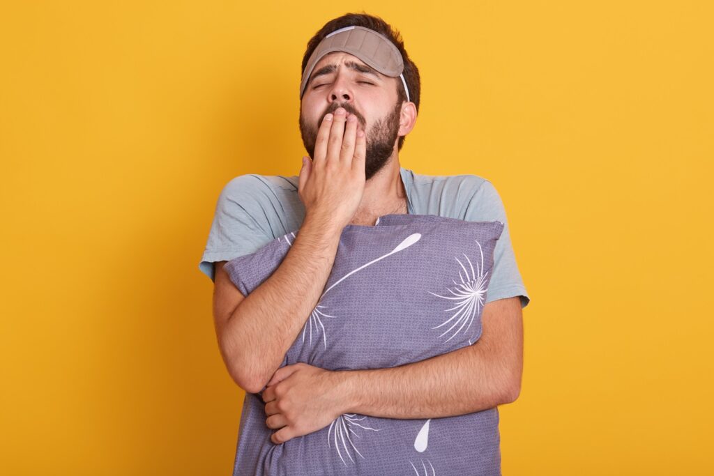 portrait sleepy young man with his pillow hands yawning covering his mouth with palms posing with blindfold forehead