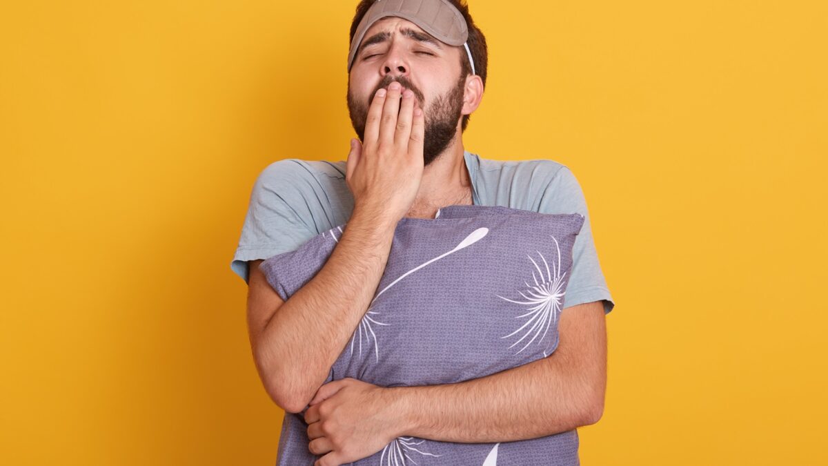 portrait sleepy young man with his pillow hands yawning covering his mouth with palms posing with blindfold forehead